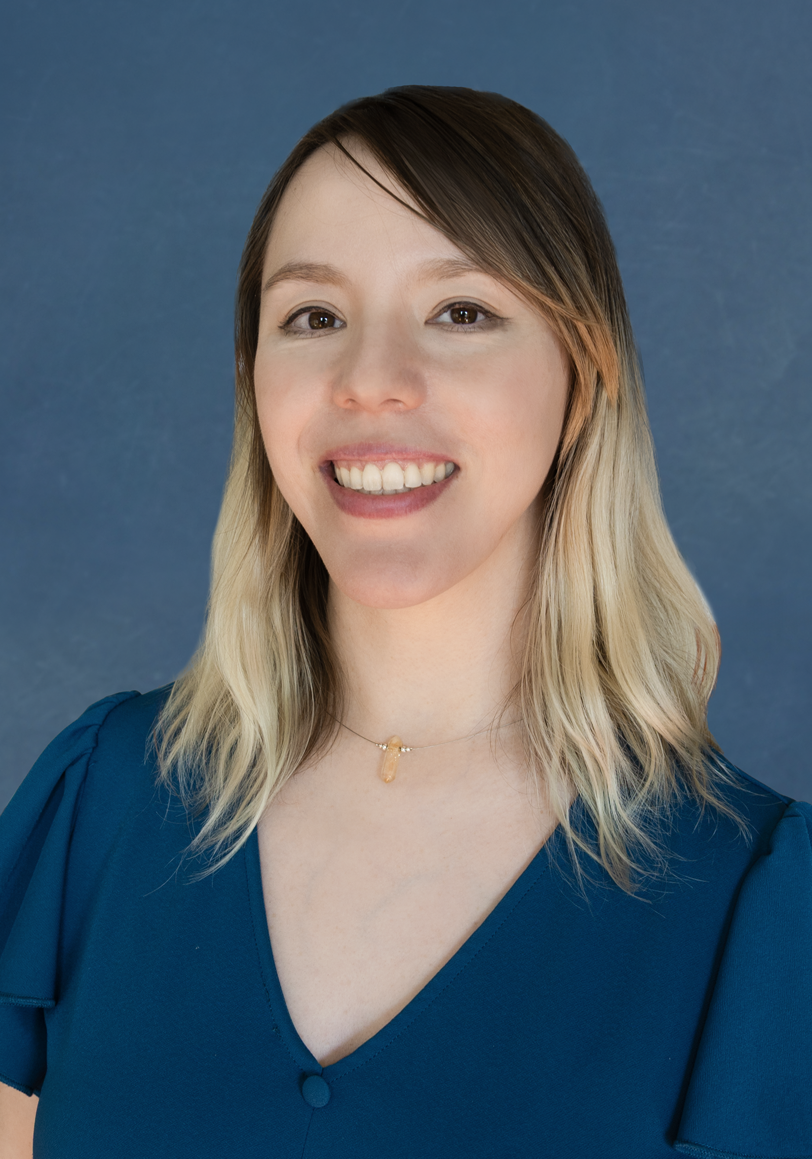 Amanda Williams Headshot Web A smiling woman with shoulder-length hair, wearing a teal blouse and a delicate necklace, poses against a blue background.